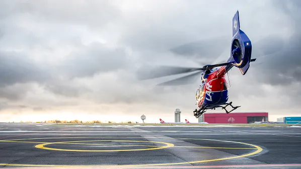 Rescue helicopter with emergency markings hovering above a helipad on an airport runway under a cloudy sky.