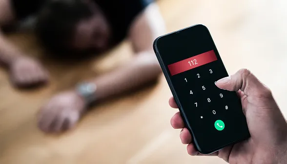 Hand holding smartphone dialling emergency number 112 with blurred person lying unconscious on the floor in the background.