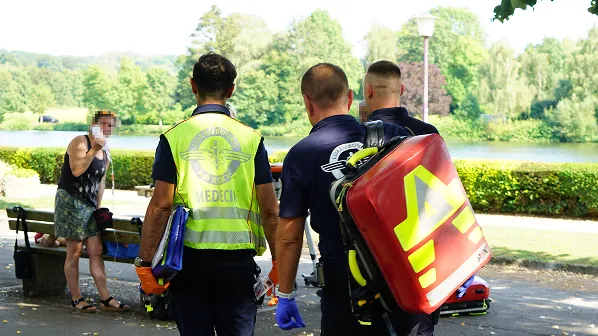 Three emergency responders, including a medic, walking towards a woman sitting on a bench near a lake in a park.