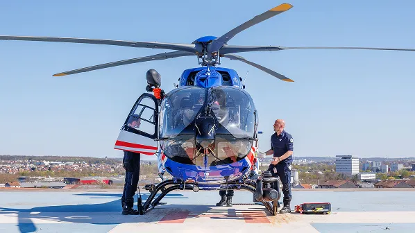 Two emergency responders preparing a blue and red helicopter on a rooftop helipad with a cityscape in the background.