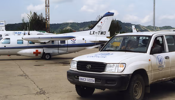 White Toyota vehicle with UNHCR logos parked on an airstrip near a small medical airplane with a red cross on the wing and tail number LX-TWO.