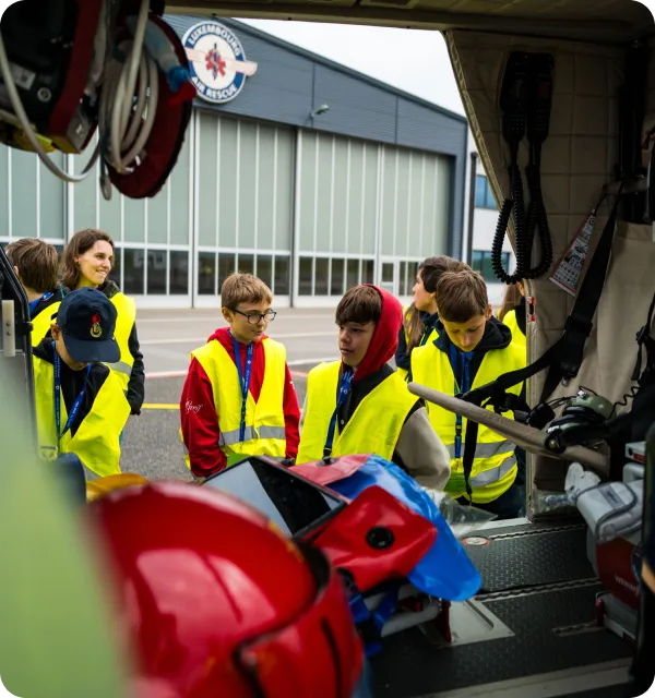 Group of children wearing yellow safety vests visiting Luxembourg Air Rescue, with fire helmets and equipment visible in the foreground.