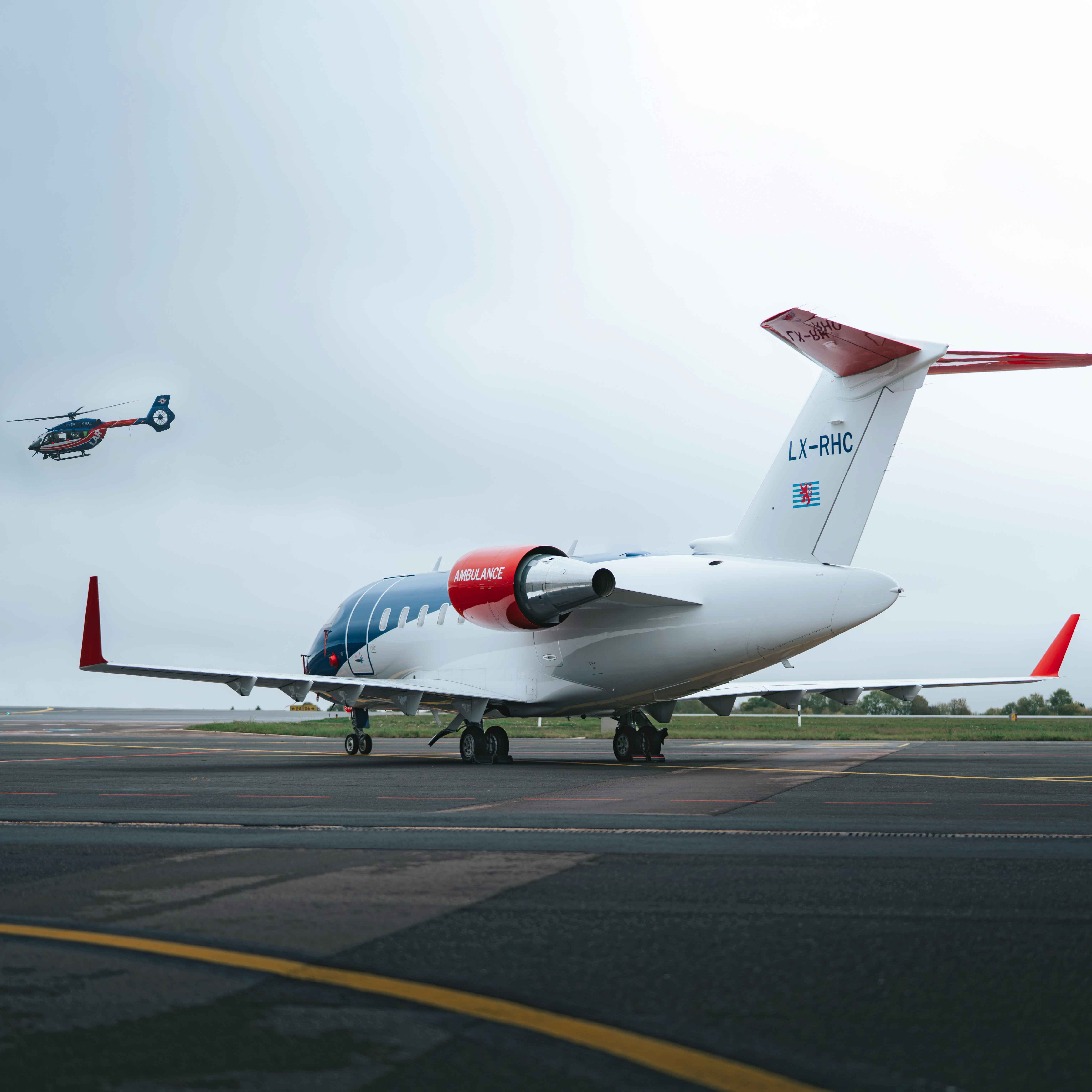 Air ambulance jet plane parked on tarmac with a helicopter flying nearby on a cloudy day.