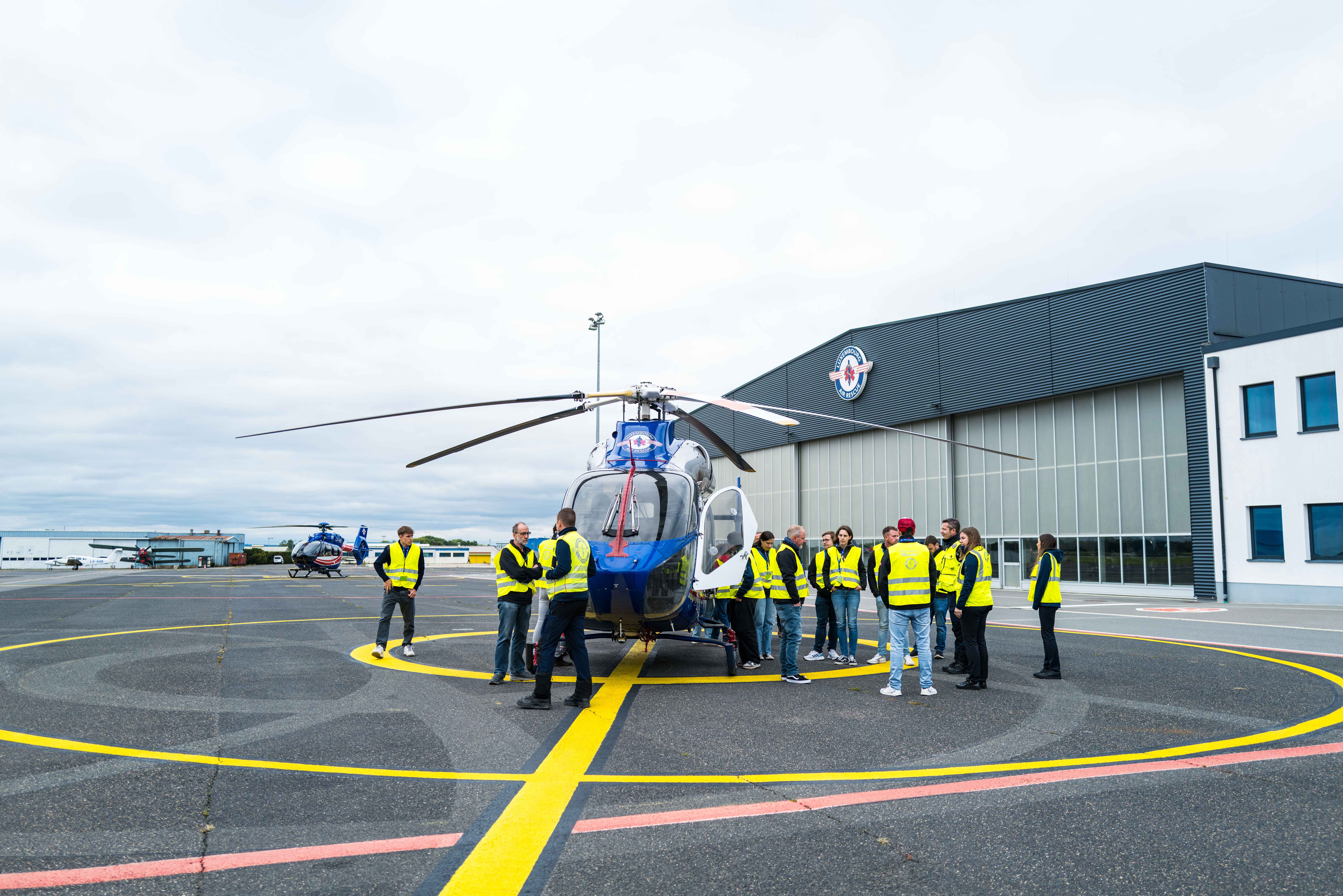 Group of people wearing yellow safety vests standing around a blue helicopter on a marked helipad outside a large hangar.