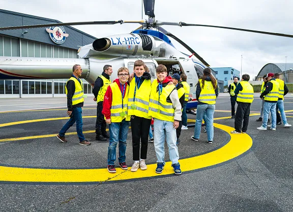 Group of people, including three children wearing yellow vests, standing on a helipad beside a helicopter.