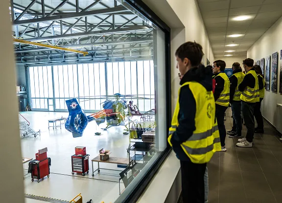Group of kids wearing yellow safety vests observing a helicopter inside a hangar through a large window.