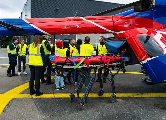Group of people wearing yellow safety vests standing near a red and blue helicopter with a stretcher on a tarmac.