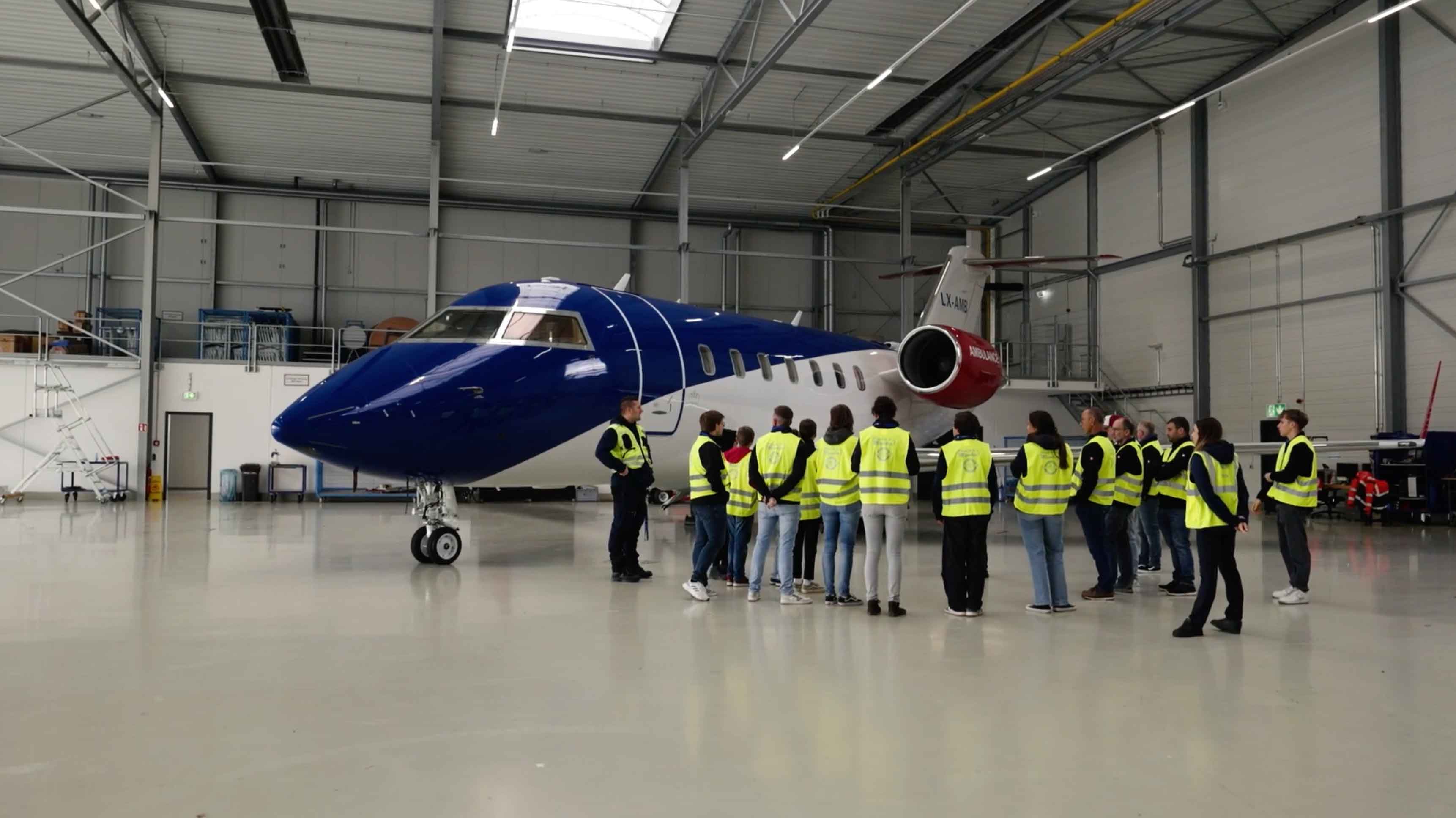 Group of people in yellow high-visibility vests receiving a guided tour next to a blue and white private jet inside an aircraft hangar.