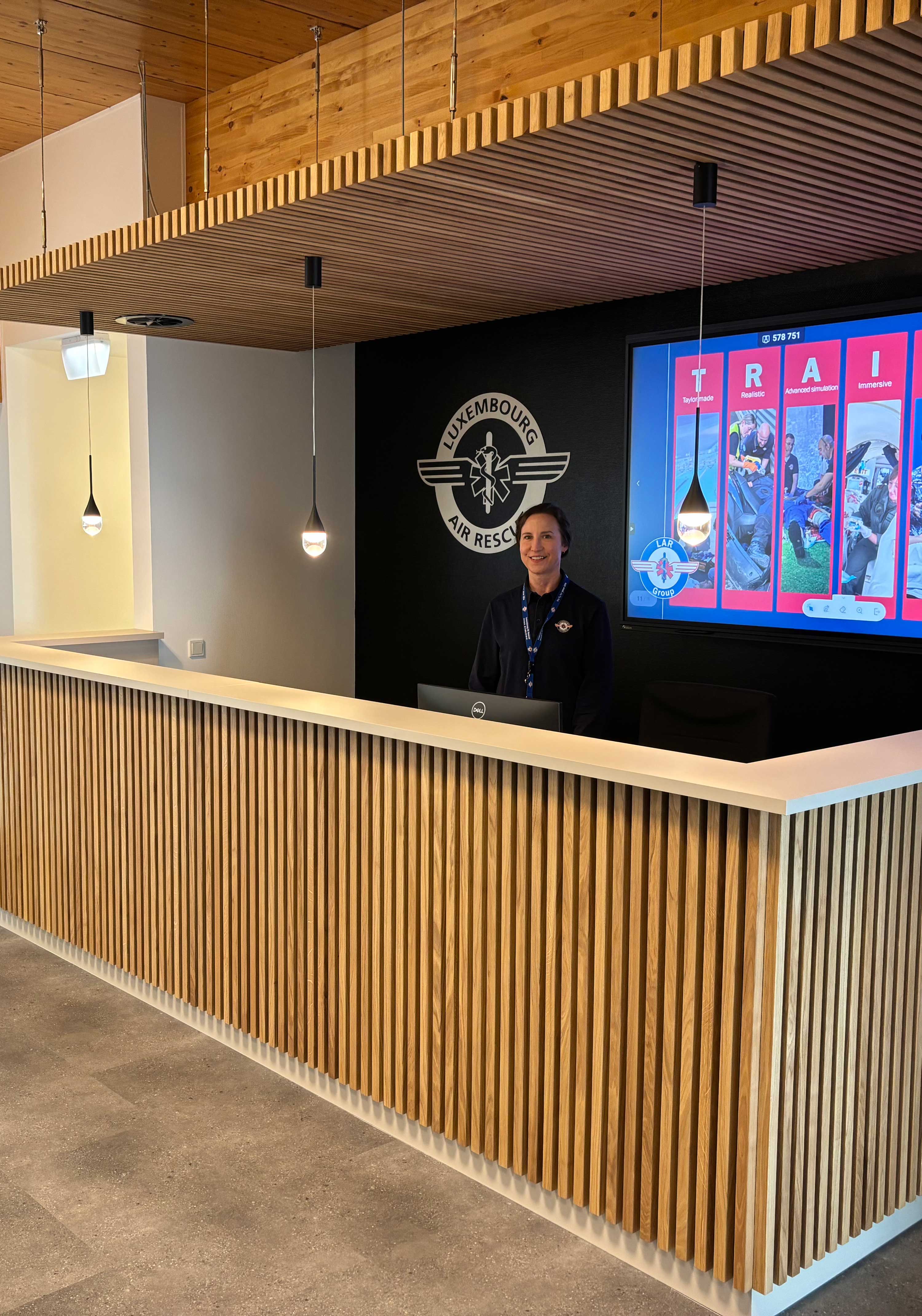 Bureau de réception avec lambris en bois, une femme souriante debout derrière, le logo du Luxembourg Air Rescue sur un mur noir et un grand écran affichant des images d'entraînement.