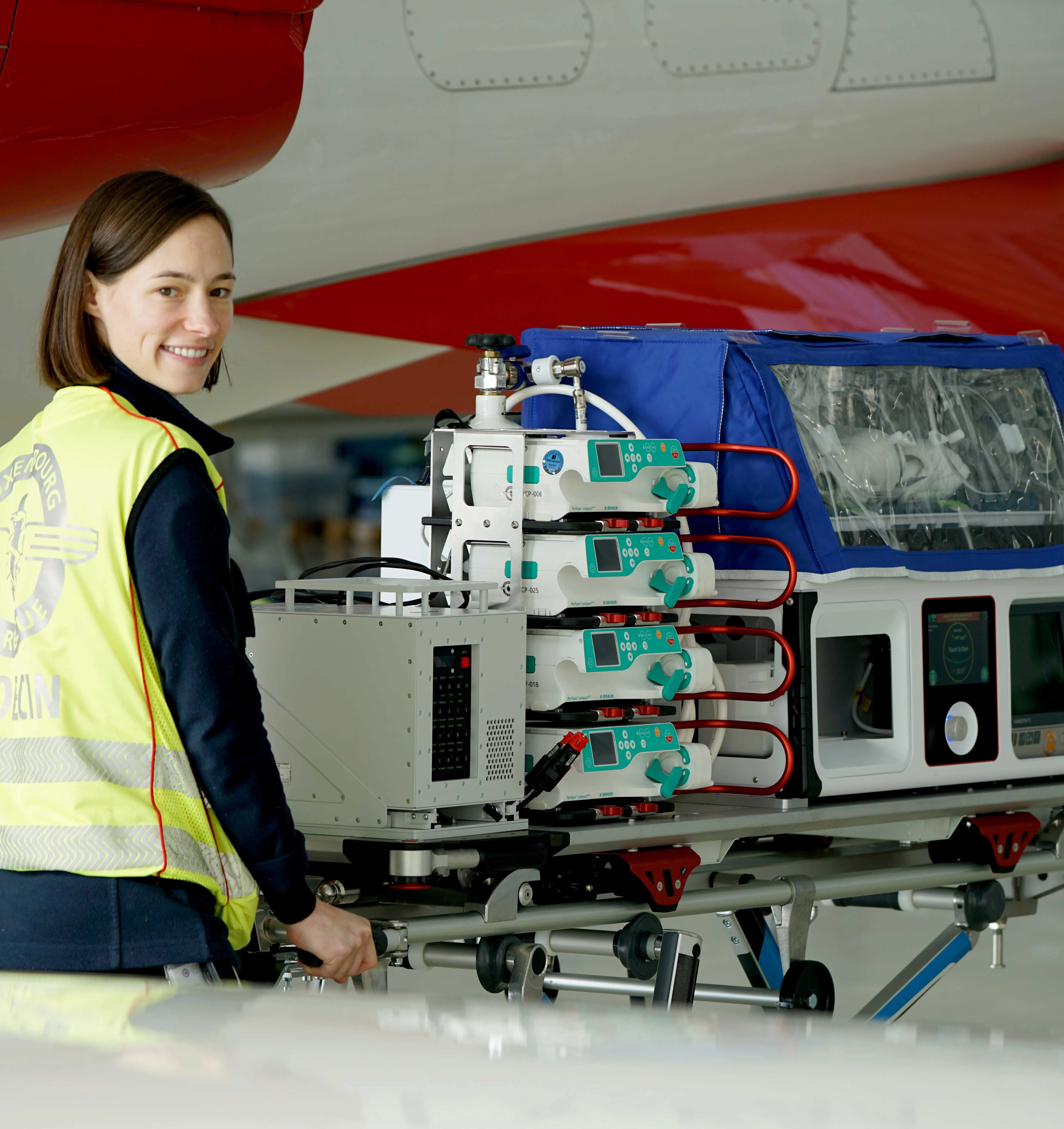 Smiling woman in a yellow reflective vest standing beside medical equipment on a stretcher under an aircraft.