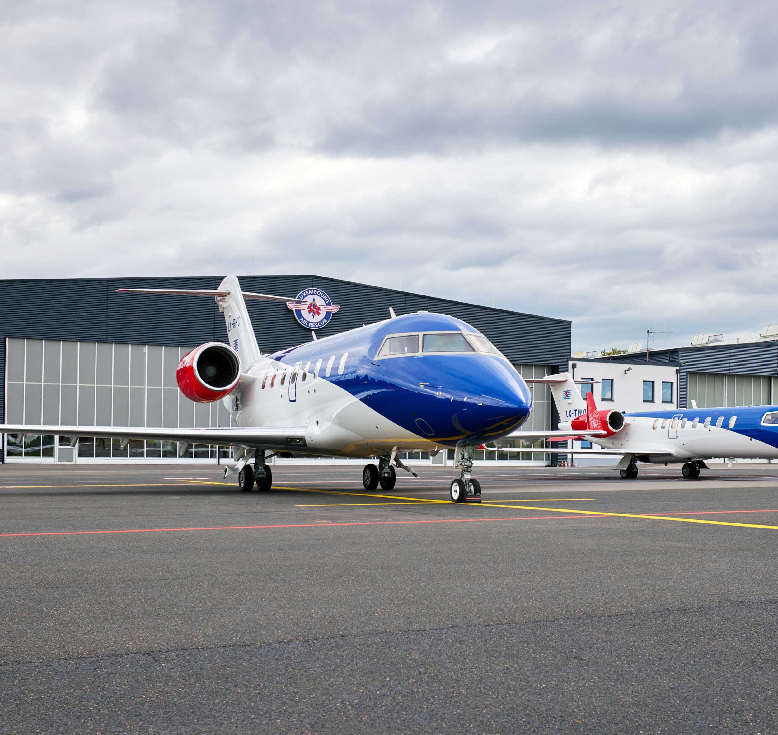 Two blue and white Luxembourg Air Rescue jets parked on an airfield in front of a hangar under a cloudy sky.