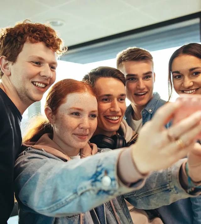 Gruppe von fünf lächelnden jungen Erwachsenen, die gemeinsam drinnen ein Selfie machen.