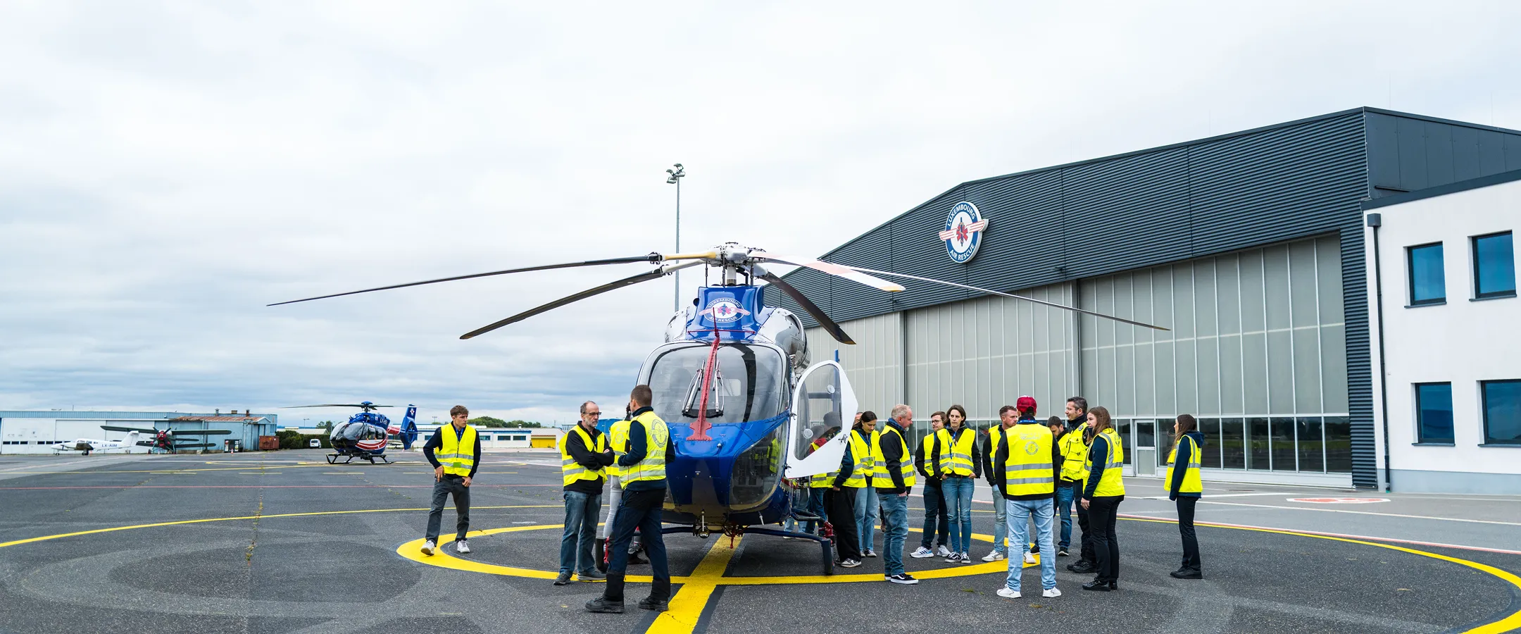 Gruppe von Personen in gelben Sicherheitswesten, die auf einem markierten Hubschrauberlandeplatz vor einem großen Hangar um einen blauen Hubschrauber stehen.