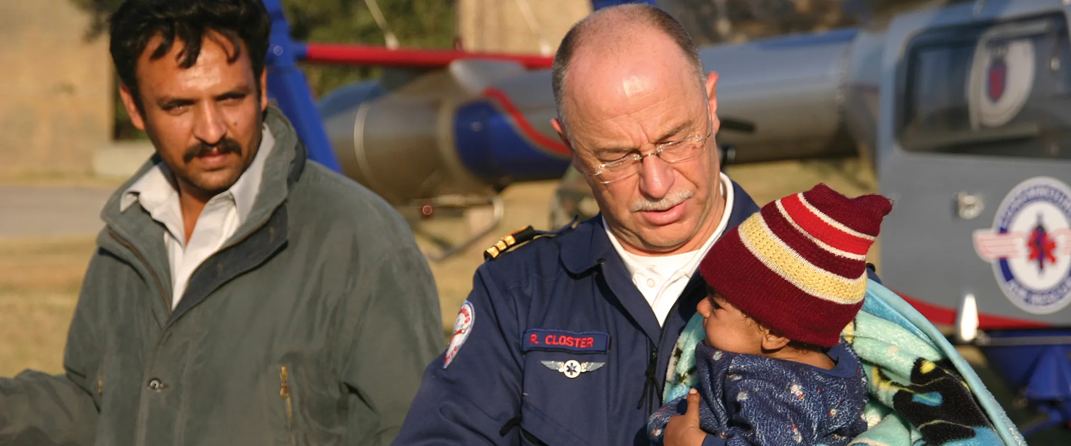 Homme en veste bleue tenant un petit enfant enveloppé dans une couverture et portant un chapeau rayé rouge, debout à côté d'un autre homme en veste grise avec un hélicoptère en arrière-plan.