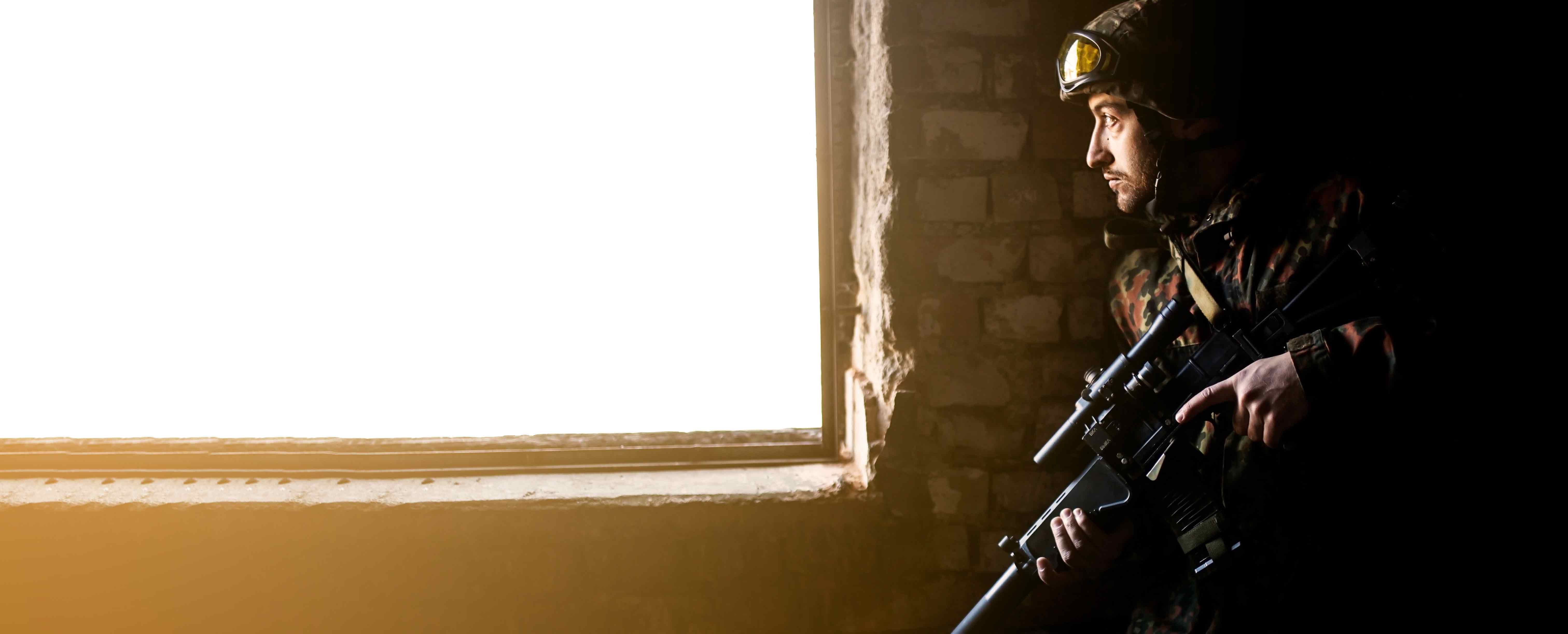 Soldat équipé en tenue camouflage observant par une fenêtre, tenant une arme avec lunette de visée.