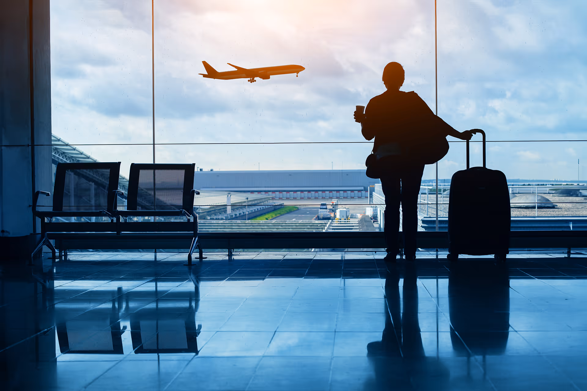 A woman stood in a departure lounge with a coffee and a suitcase watching a plane taking off