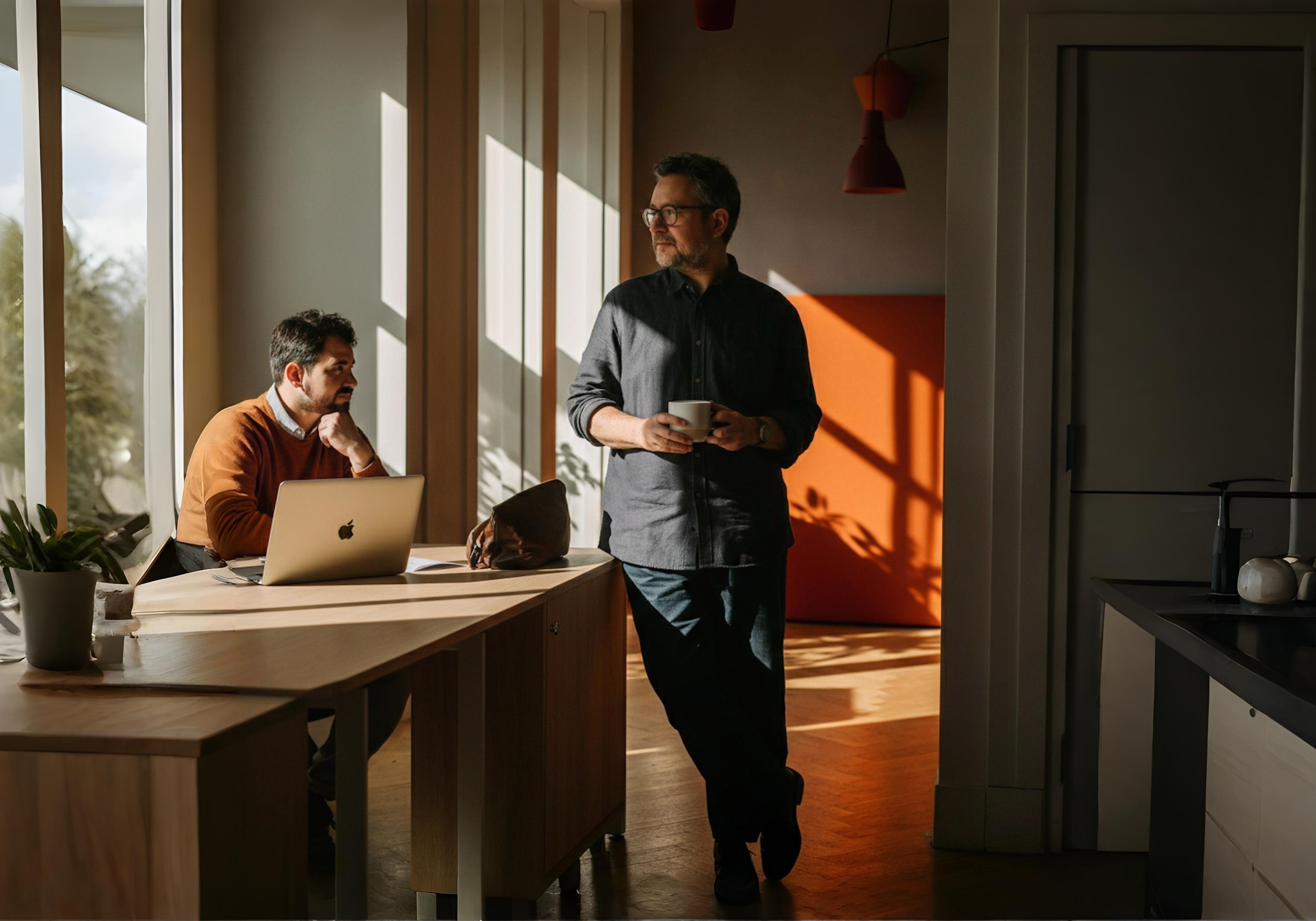 Two men in a sunlit modern office; one sits at a desk with a laptop, the other stands holding a cup.