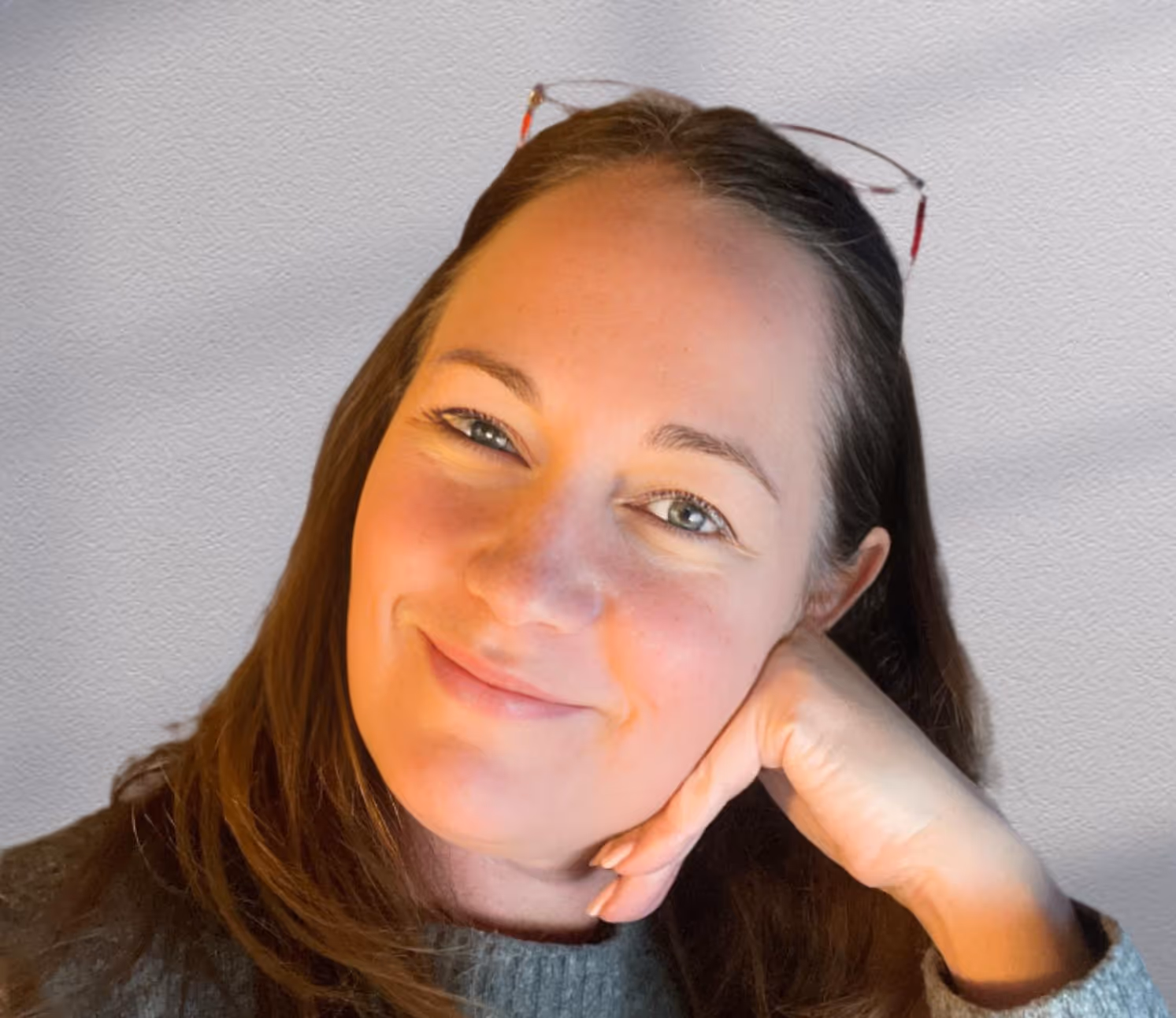Smiling woman with long brown hair resting her head on her hand, wearing a gray sweater and glasses on her head.