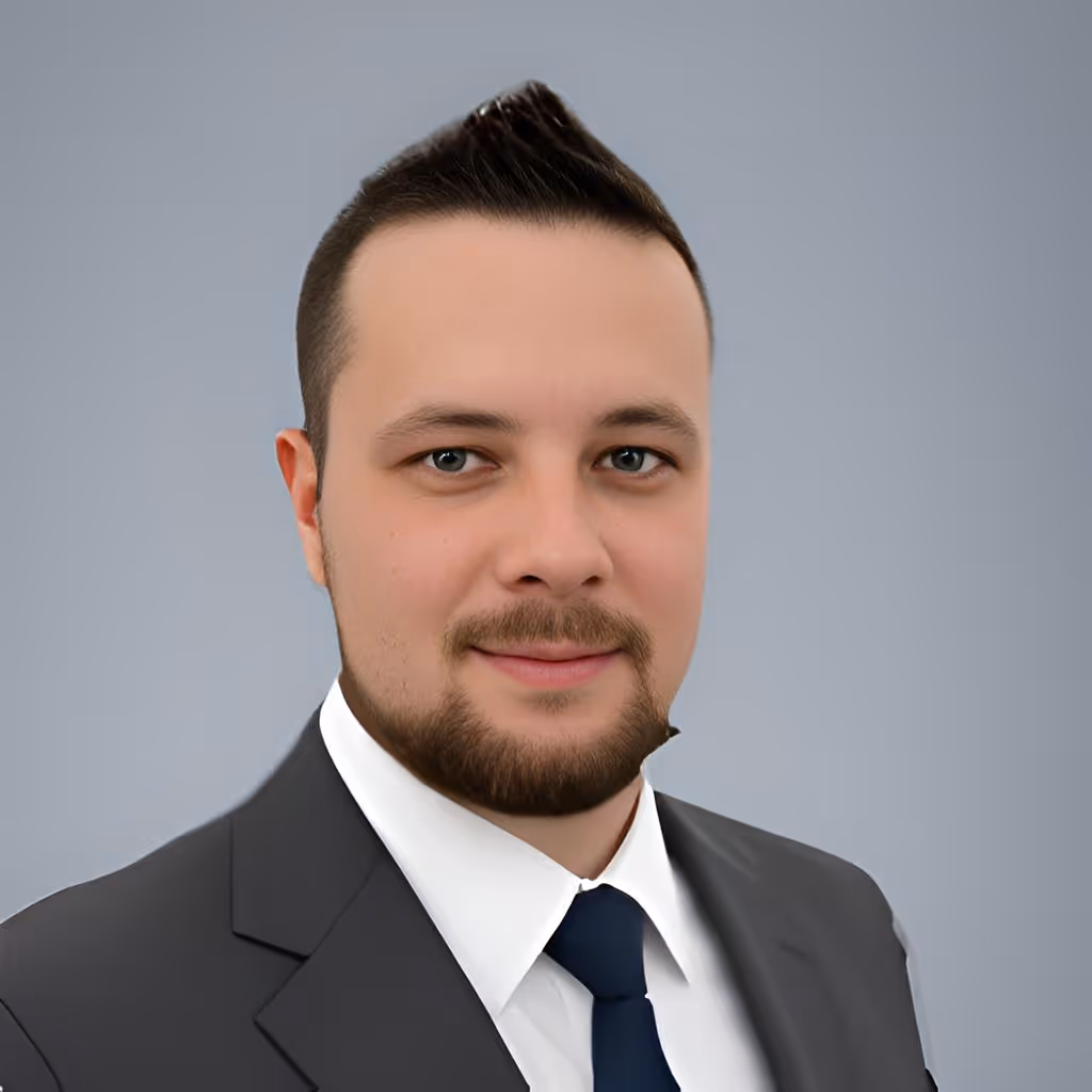Young man with short dark hair and beard wearing a dark suit, white shirt, and navy tie with a neutral gray background.