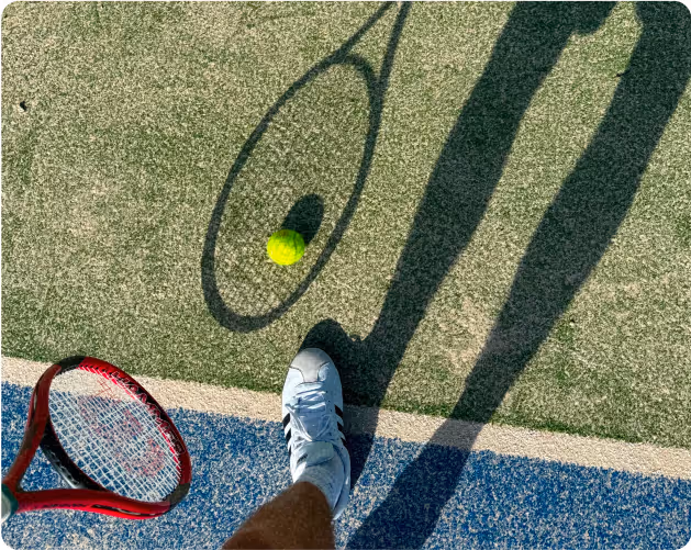 Tennis racket, ball, and player's shoe on a court with long shadows