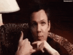 A man in a patterned armchair makes air quotes.