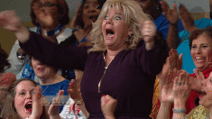 A woman in a crowd with blonde hair reacts with a shocked, excited expression, throwing her fists in the air.