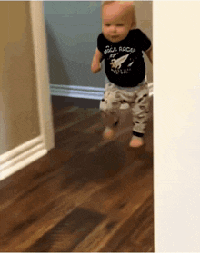 A baby in a black t-shirt and patterned pants walks down a hallway.