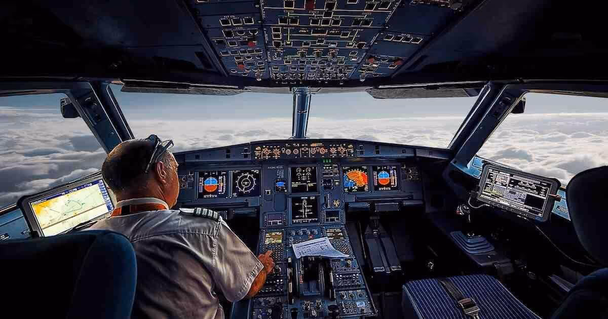 A pilot sits at the controls of an airplane cockpit, high above a cloud layer.