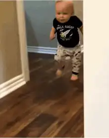 A baby in a black t-shirt and patterned pants walks down a hallway.