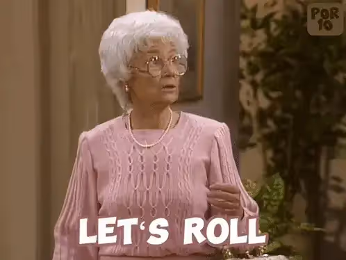 A woman with white hair and glasses, wearing a pink sweater, with the text "LET’S ROLL" on screen
