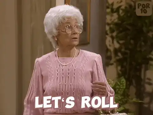 A woman with white hair and glasses, wearing a pink sweater, with the text "LET’S ROLL" on screen