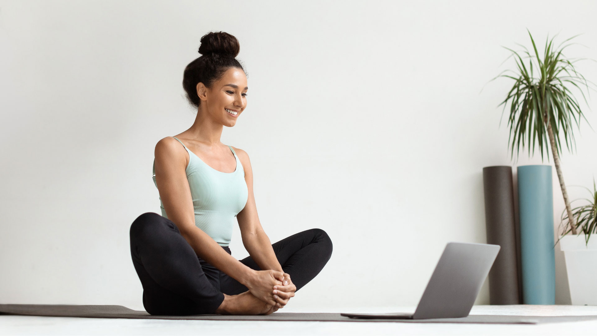 A smiling woman sitting on a yoga mat in butterfly pose, looking at her laptop during an online yoga class in a bright home studio.