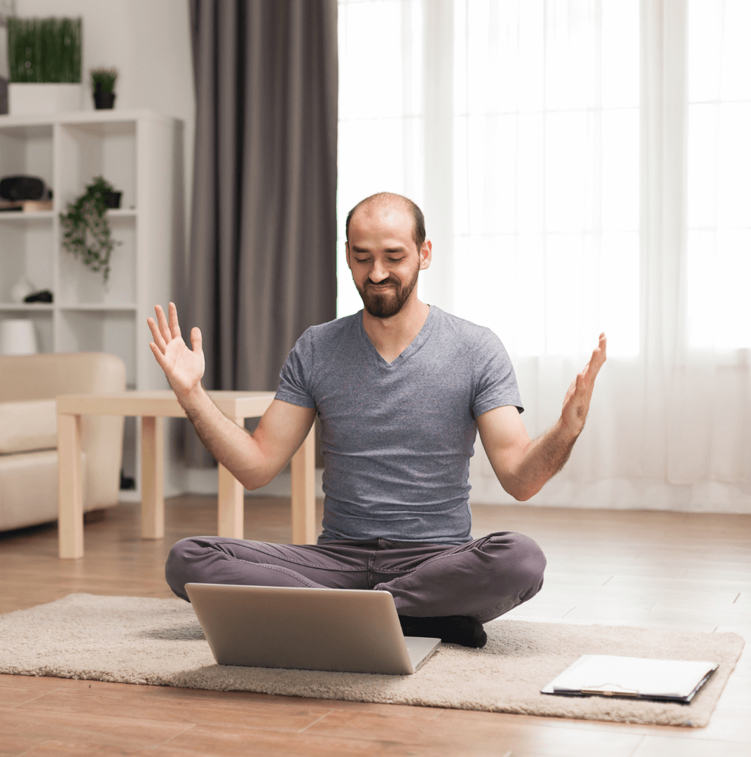 A man sits cross-legged on a rug with a laptop, his arms raised and eyes closed.