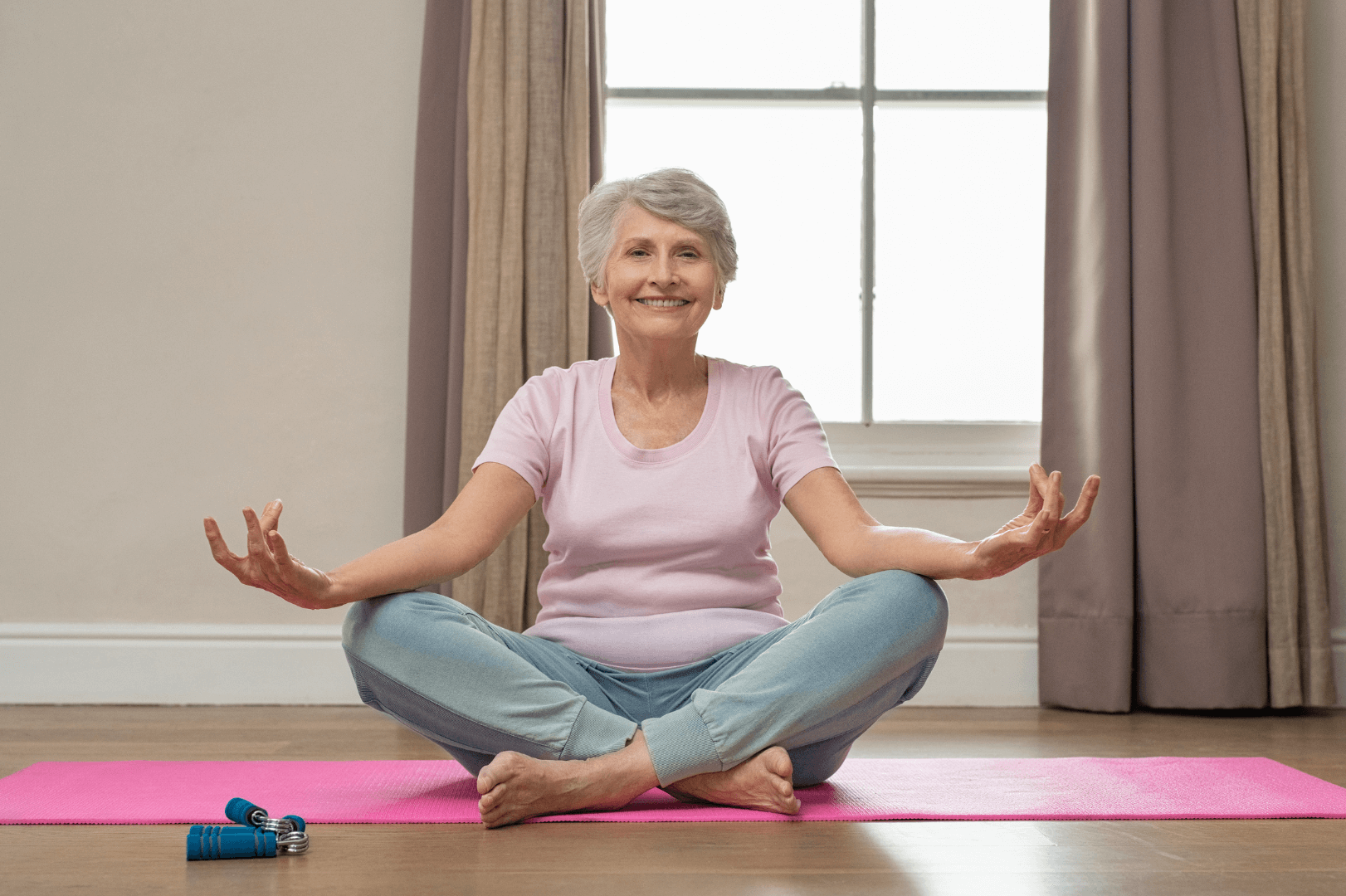 A happy senior woman sits cross-legged on a pink yoga mat with her hands in a meditation gesture.