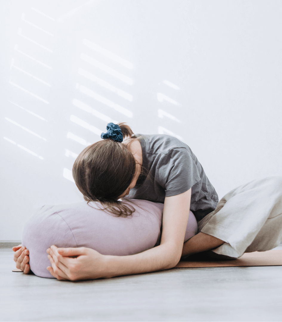 A woman uses a bolster to rest her head during a seated forward bend ina  restorative yoga session.