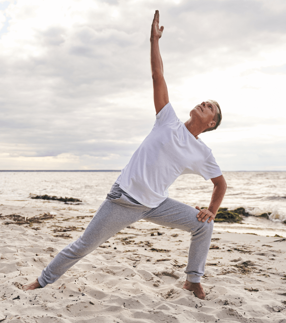 A man in a white t-shirt and gray sweatpants does a reverse warrior pose on a sandy beach.