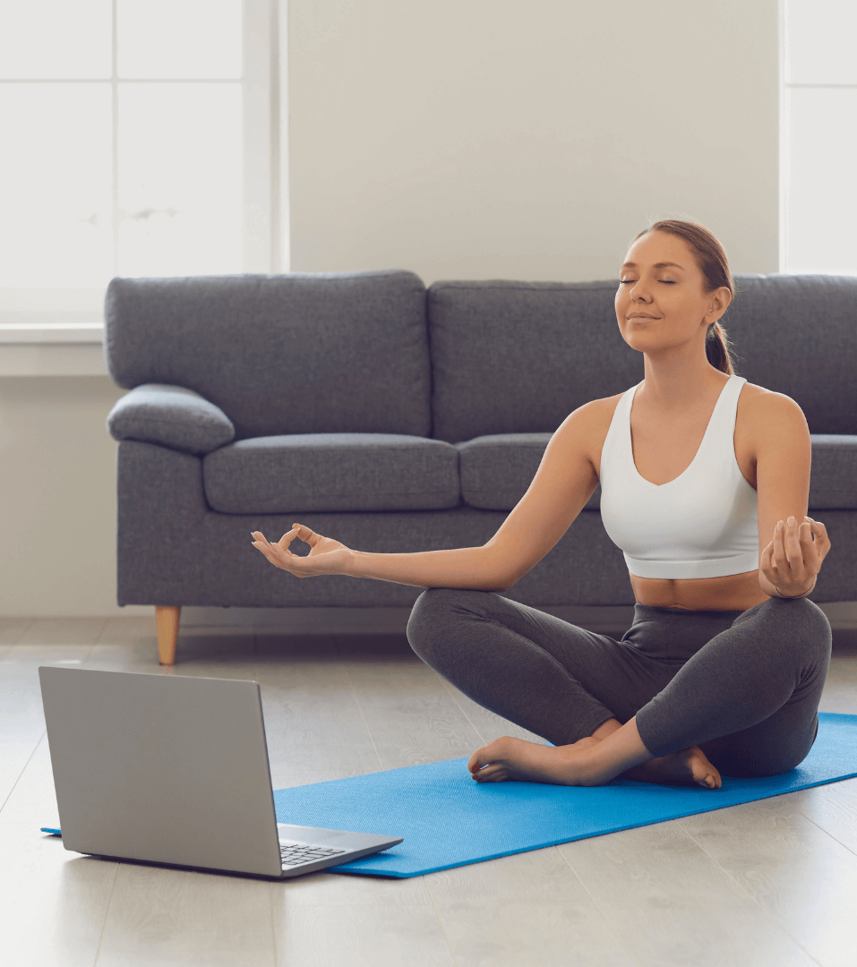 A woman sits in a meditation pose on a blue mat with a laptop open in front of her.