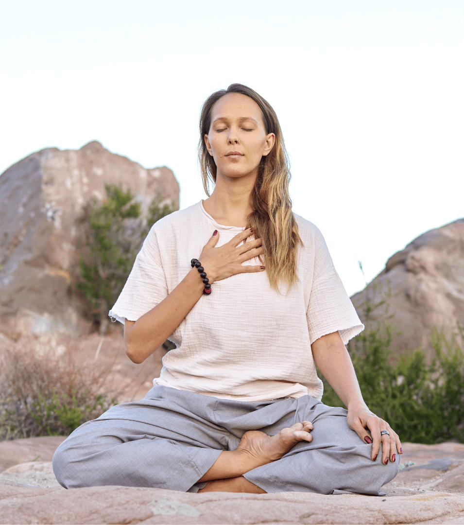 A woman meditates in a cross-legged position on a rock, with her eyes closed and one hand on her chest.