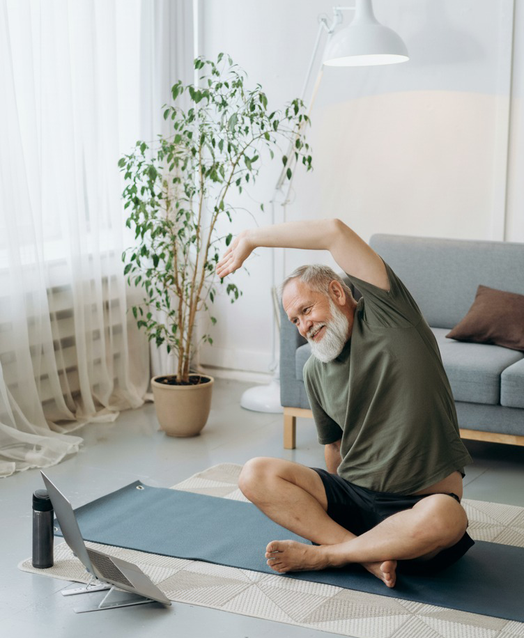 A man with a white beard sits in a cross-legged position on a mat, meditating with his eyes closed.