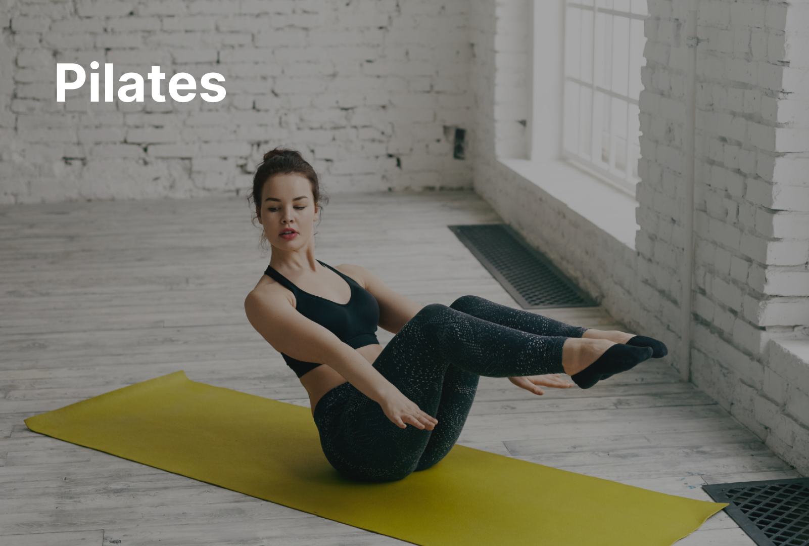 A woman with short, blonde hair in a white sports bra and gray leggings lies on her stomach on a black mat. She is holding a sphinx yoga pose.