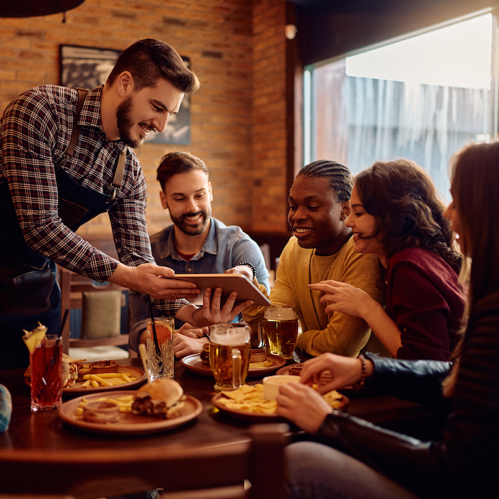A group of friends sitting at a restaurant looking at the iPad the server is presenting to them.