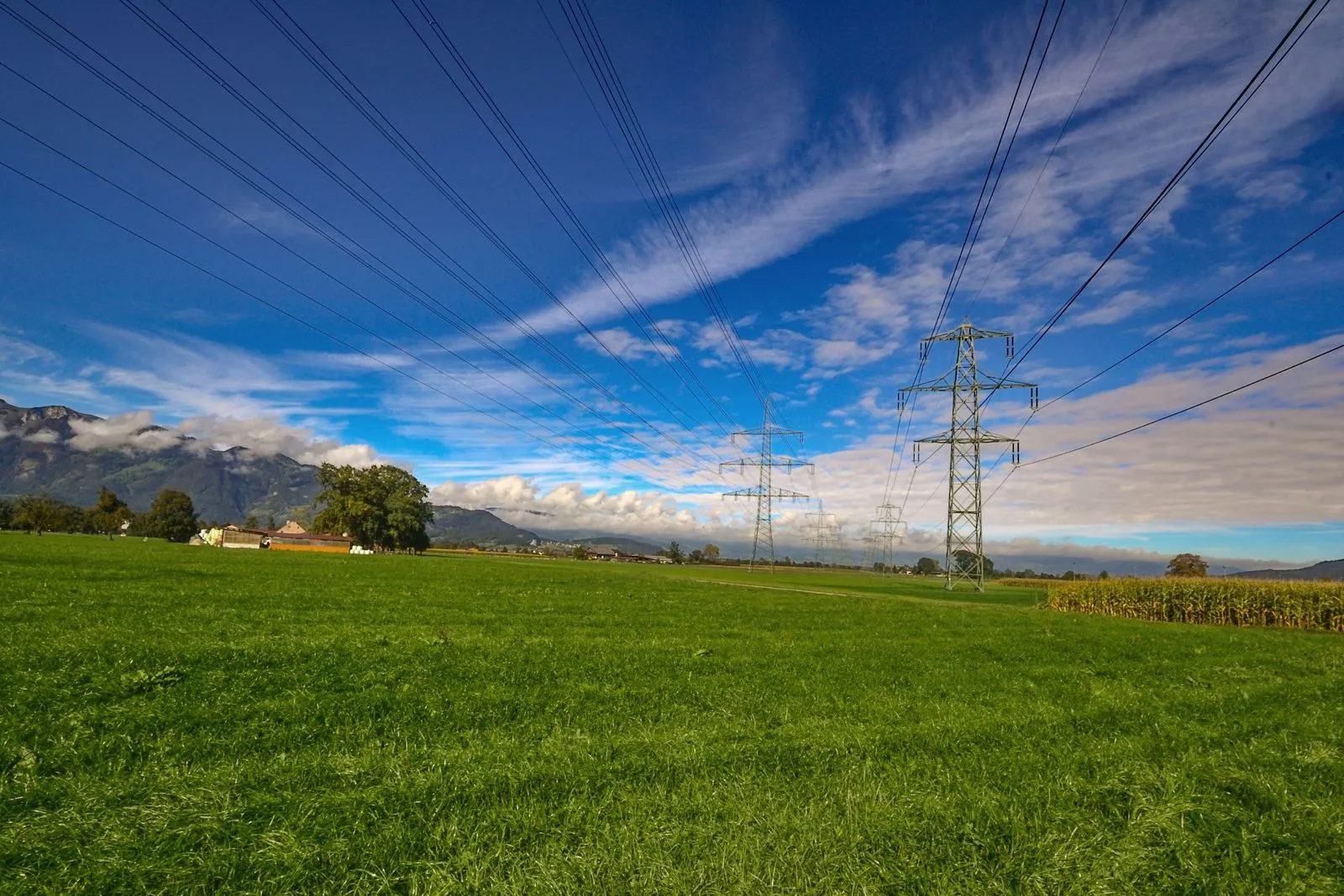 High-voltage power lines crossing a green field with mountains under a blue sky