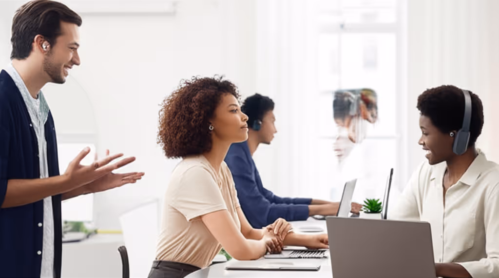 People are working at computers in an office. A man is clapping and two wear headphones.
