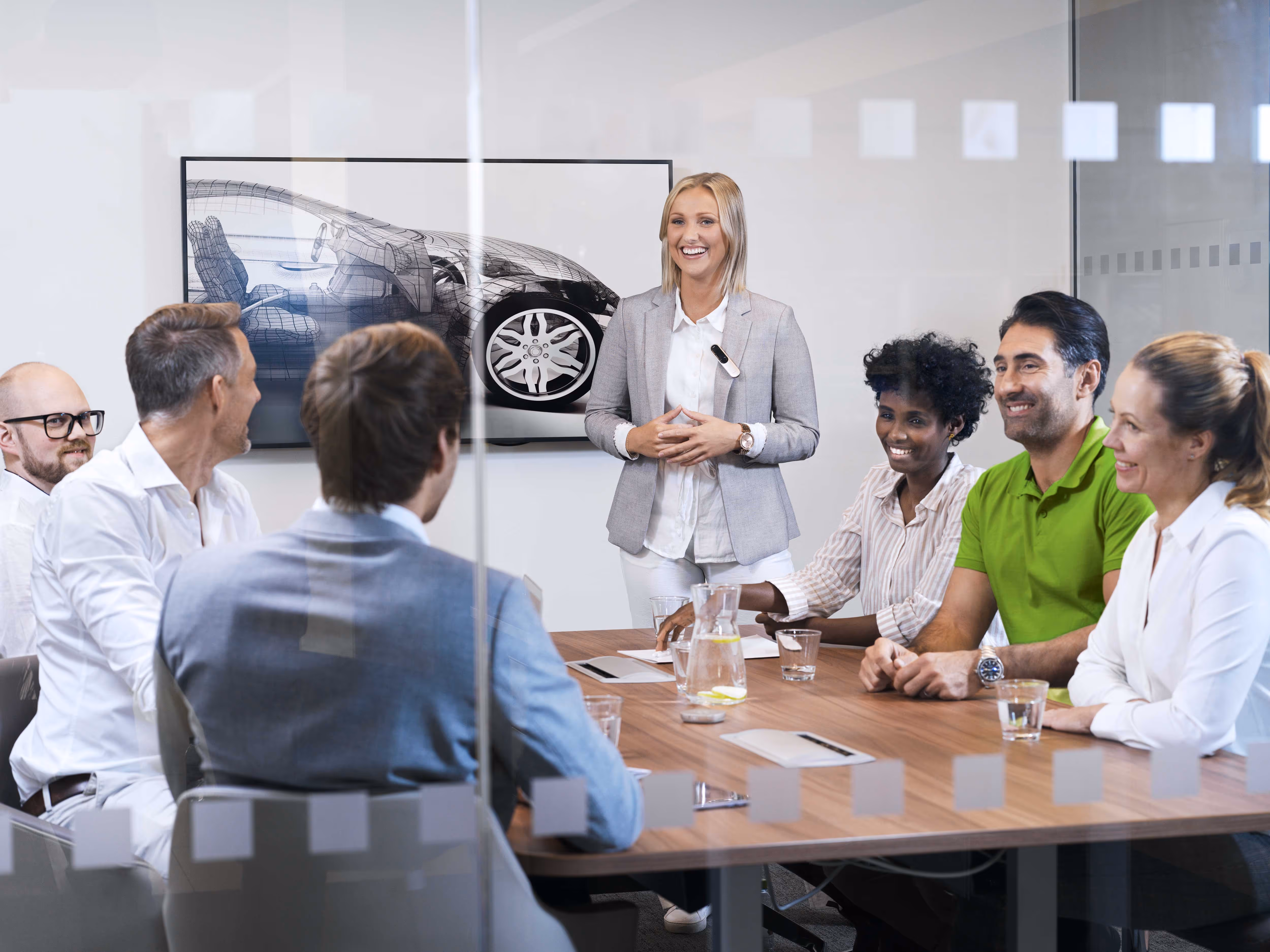 A woman stands and presents to a group of six seated people in a conference room.