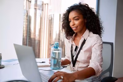 Woman sitting at a desk, using a laptop and holding a water bottle, with cityscape background.