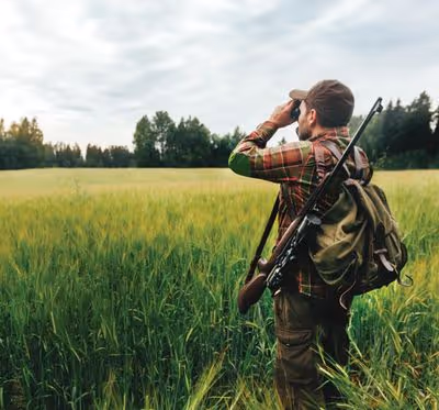 A person in a field is looking through binoculars, with a rifle and backpack.