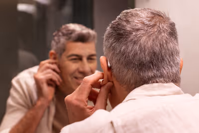 Man putting on a hearing aid while looking into a bathroom mirror.