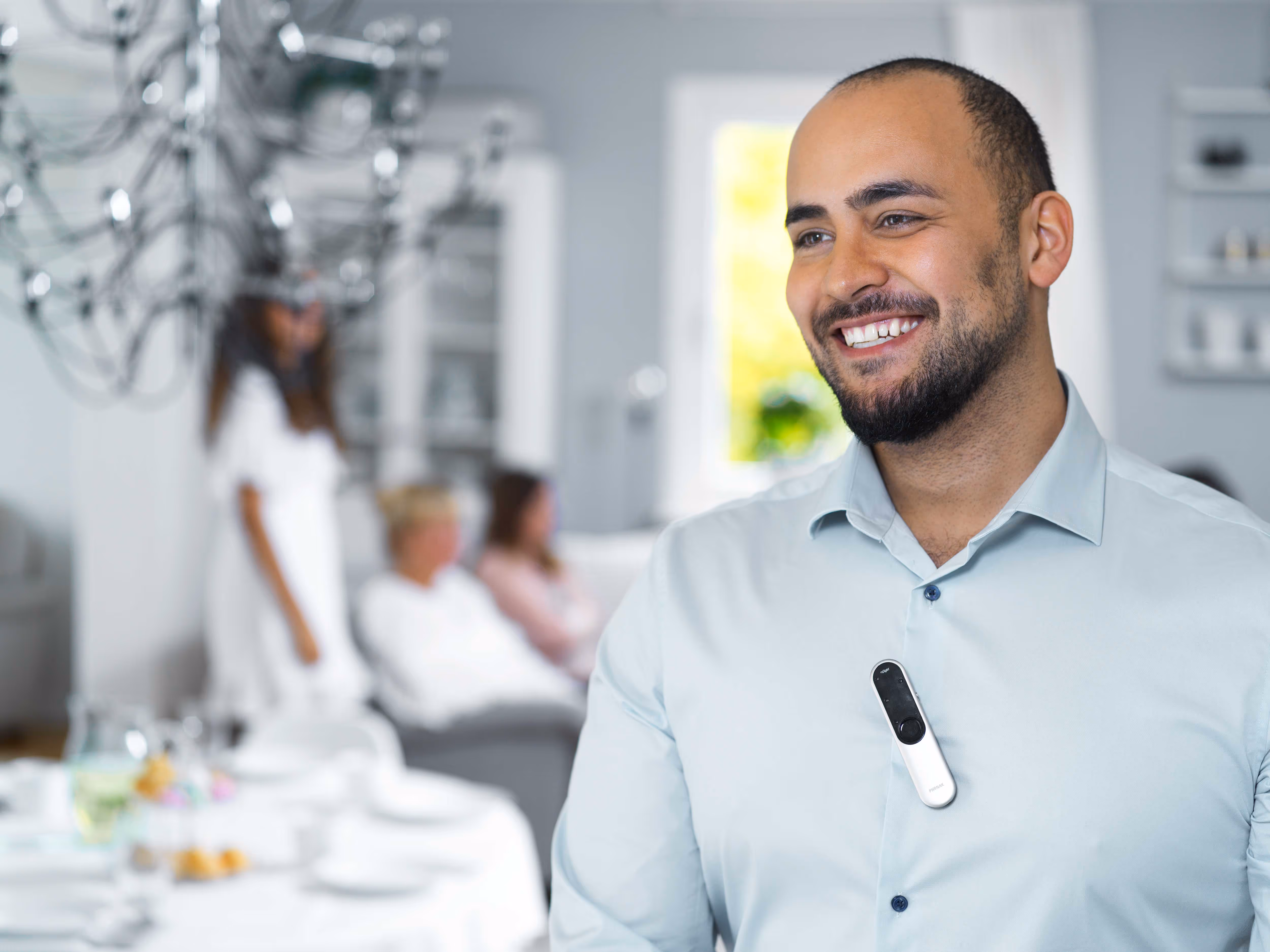 A man with a clip-on device smiles, while people sit on a couch in the background.