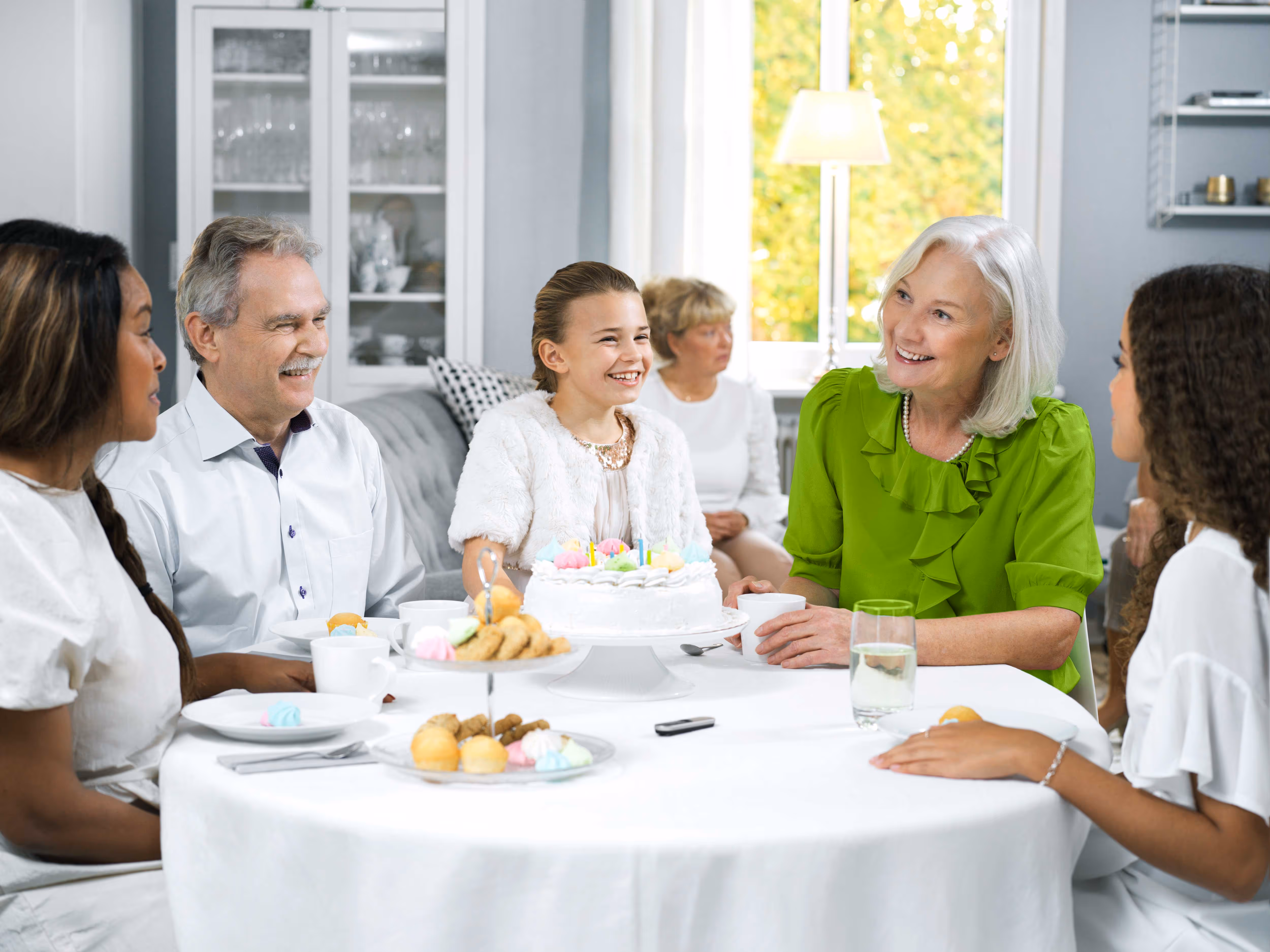 A group of people sitting around a table with a cake, smiling and talking together.