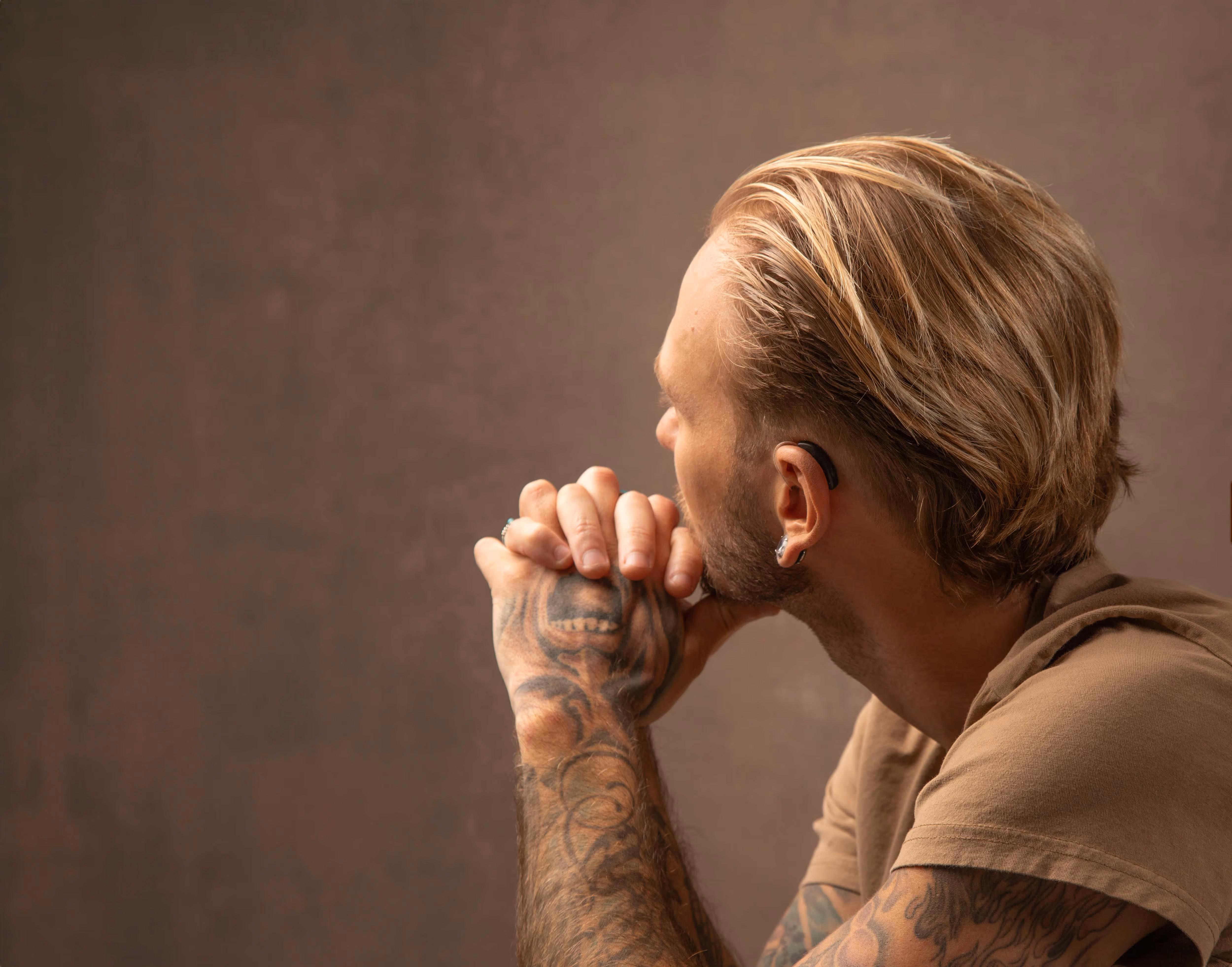 A person with tattoos and blond hair is sitting, looking off to the side.